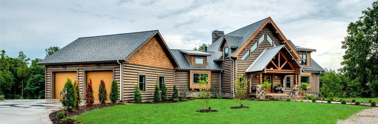 Photo of the front of the Patterson with double a-frame front entrance with the upper level containing geometric windows. an enclosed breezeway attaches the two-car garage to the home The round logs provide an architectural design element and chink is low profile